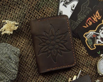 May include: A dark brown leather wallet with an embossed edelweiss flower design. The wallet is rectangular and has visible stitching. Other items include patches with graphics and a vintage stopwatch.