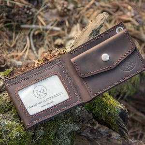 May include: A brown leather wallet with a stitched design and a small pocket on the outside. The wallet has a clear window with the text "Handmade Leather Goods from Ukraine".