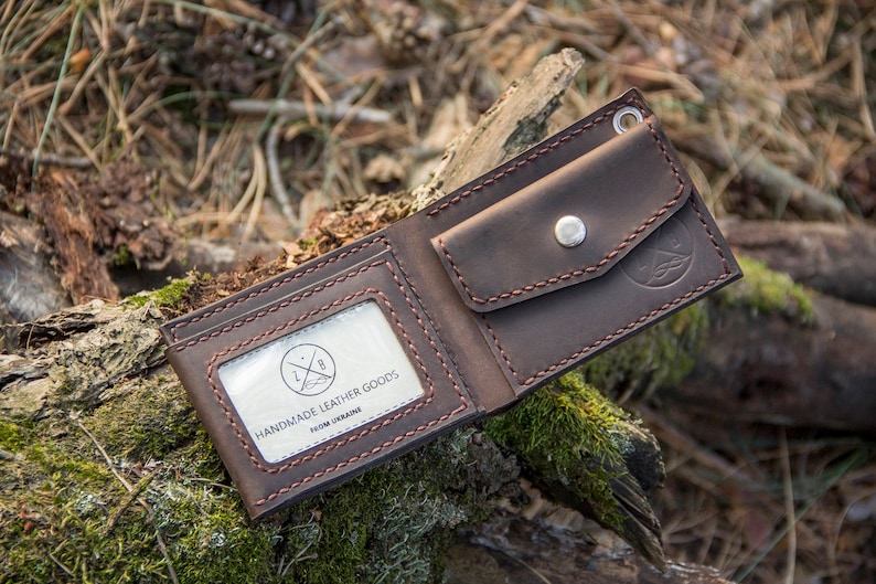 May include: A brown leather wallet with a stitched design and a small pocket on the outside. The wallet has a clear window with the text "Handmade Leather Goods from Ukraine".