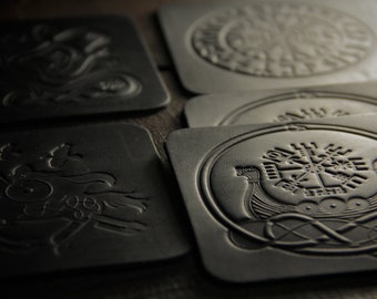 May include: Four black leather coasters with embossed designs. The coasters feature a variety of Norse symbols and imagery, including a ship, a compass, and a wolf.