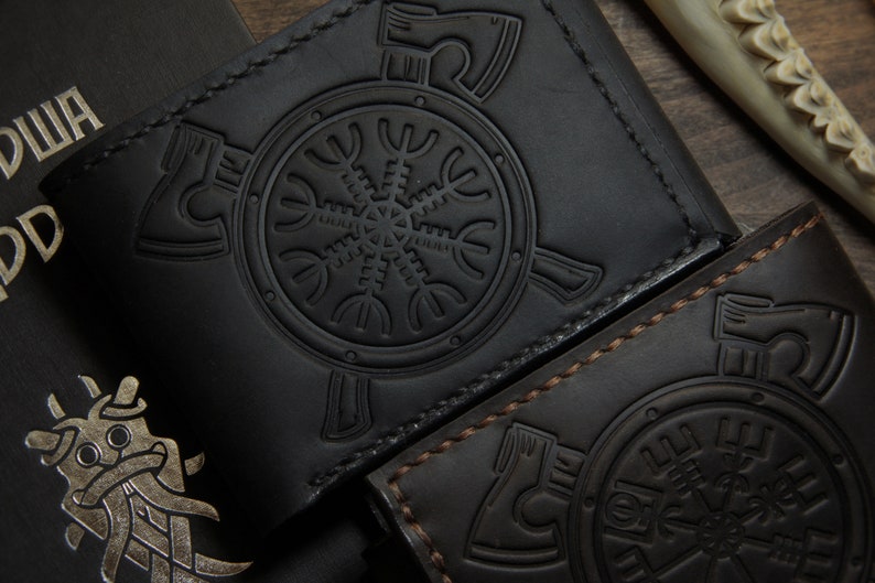 May include: Two black leather wallets with embossed Viking-style designs. One wallet is black with a matching black stitch, and the other is dark brown with a contrasting brown stitch. The wallets feature a circular emblem with axes and a central symbol.