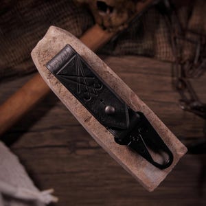 May include: A black leather keychain strap featuring an embossed sigil design and a silver-toned metal snap button. The strap is attached to a black metal lobster clasp, providing a secure attachment point for keys or other small personal items.
