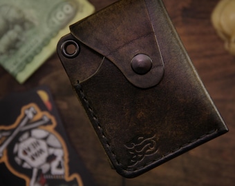 May include: Dark brown leather wallet with a flap closure and a metal snap. The wallet has a stitched border and a decorative emblem on the front. A hundred-dollar bill and a skull are in the background.