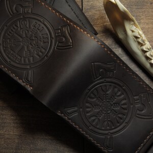 May include: Dark brown leather wallet with embossed Viking symbols. The wallet features a detailed design with stitched edges. The background includes a wooden surface and a bone.