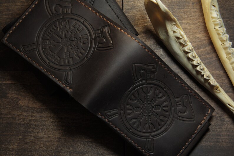 May include: Dark brown leather wallet with embossed Viking symbols. The wallet features a detailed design with stitched edges. The background includes a wooden surface and a bone.