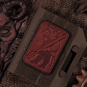 May include: A rectangular dark brown leather patch featuring an embossed illustration of a stylized warrior figure holding a spear. The patch is stitched onto a dark olive green fabric base with a hook-and-loop fastener backing for attachment.