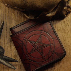 Puede incluir: Una cartera de cuero roja con un diseño de pentagrama y una cabeza de cabra en el centro. La cartera está cerrada y tiene un borde cosido.
