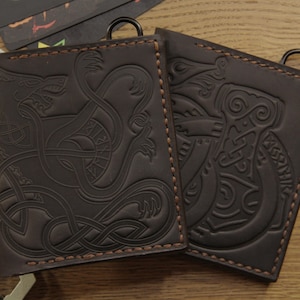 Puede incluir: Dos carteras de cuero marrón oscuro con intrincados diseños en relieve. Una presenta un dragón, la otra un martillo. Ambas tienen un anillo de metal y están cosidas con hilo marrón claro. Las carteras están sobre una superficie de madera.
