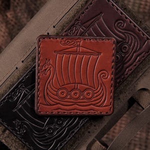 May include: A square, reddish-brown leather patch featuring an embossed Viking longship design with a dragon-headed prow and a striped sail. The patch is stitched along the edges and is positioned over a dark brown leather piece and a tan fabric base.