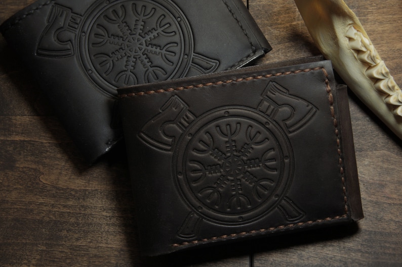 May include: Two leather wallets, one black and one brown, are displayed on a wooden surface. Each wallet features an embossed design of an axe and a circular symbol. The brown wallet has visible stitching.