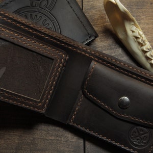 May include: Open, dark brown leather wallet with a clear ID window and a snap closure. The wallet has visible stitching and a small embossed design. A black wallet is visible in the background.
