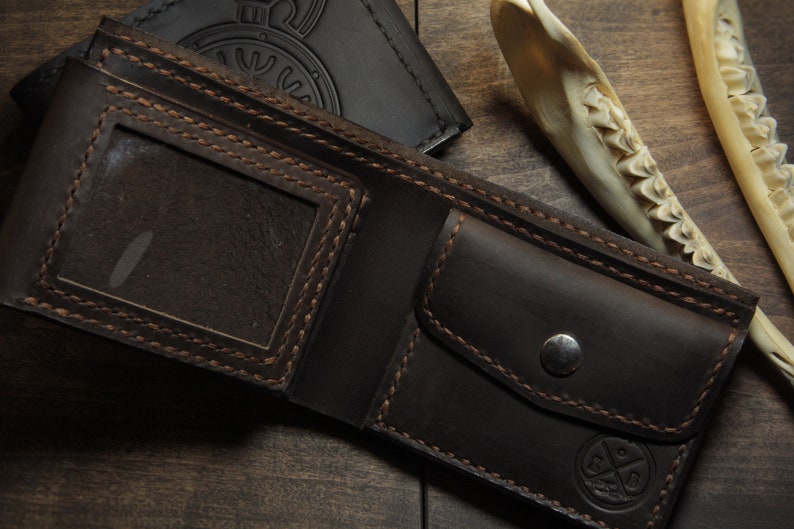 May include: Open, dark brown leather wallet with a clear ID window and a snap closure. The wallet has visible stitching and a small embossed design. A black wallet is visible in the background.