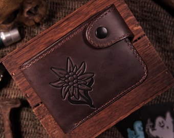 May include: A dark brown leather bifold wallet featuring a debossed edelweiss flower design on the front. The wallet is secured with a matching brown leather strap and a single black metal snap button closure, with visible stitching along the edges.