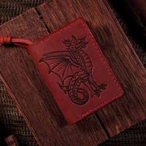 May include: A rectangular red leather passport holder featuring a debossed black dragon illustration on the front cover. The cover includes a matching red leather cord tied in a knot at the top left corner and is stitched along the edges.