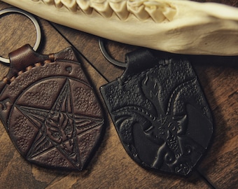 Può includere: Due portachiavi in ​​pelle con finitura marrone scuro e nero. Entrambi i portachiavi presentano un design pentagramma con una testa di capra al centro.