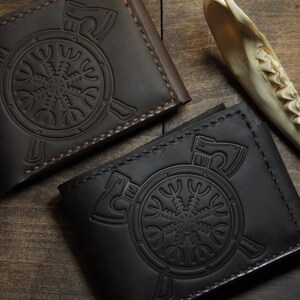 May include: Two leather wallets, one dark brown and one black, are displayed on a wooden surface. Each wallet features an embossed design of a circular emblem with axes and a stylized symbol. The wallets are closed, showcasing their detailed craftsmanship.