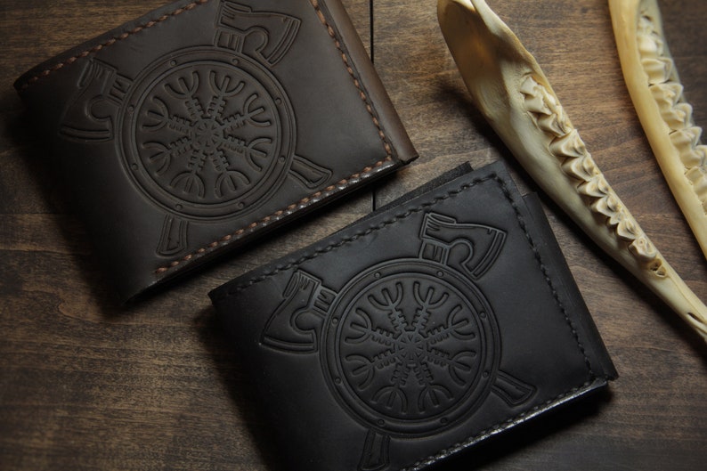 May include: Two leather wallets, one dark brown and one black, are displayed on a wooden surface. Each wallet features an embossed design of a circular emblem with axes and a stylized symbol. The wallets are closed, showcasing their detailed craftsmanship.