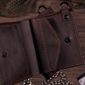 May include: Open brown leather wallet with visible stitching and a snap closure. The wallet has multiple compartments and a metal ring attachment. The leather appears to be distressed, giving it a vintage look. The wallet is on a wooden surface.