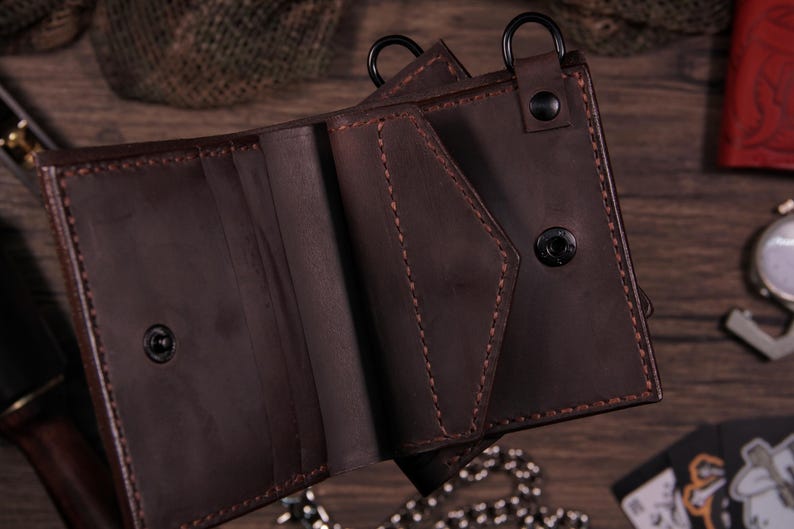 May include: Open brown leather wallet with visible stitching and a snap closure. The wallet has multiple compartments and a metal ring attachment. The leather appears to be distressed, giving it a vintage look. The wallet is on a wooden surface.