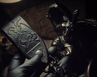 Pode incluir: Porta-chaves de couro preto com um design em relevo detalhado de uma figura demoníaca. O porta-chaves apresenta um fecho de metal e um fecho de botão. Um acessório escuro de estilo gótico.