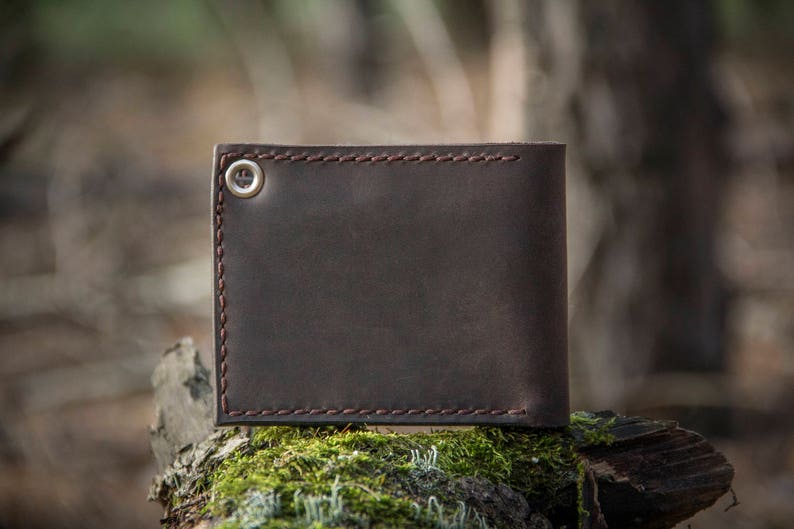 May include: A brown leather wallet with a stitched edge and a silver button. The wallet is sitting on a mossy log.