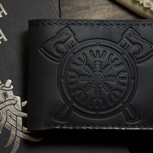 May include: Black leather wallet with embossed Viking symbols, including axes and a shield with a runic design. The wallet is next to a book with silver lettering and a stylized illustration.