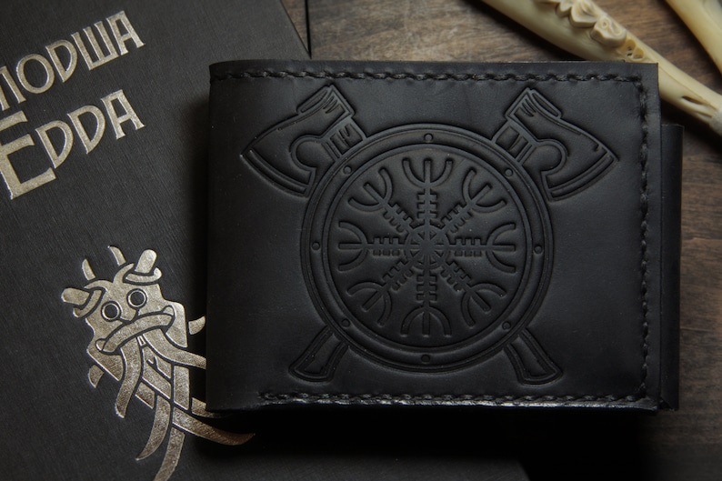 May include: Black leather wallet with embossed Viking symbols, including axes and a shield with a runic design. The wallet is next to a book with silver lettering and a stylized illustration.