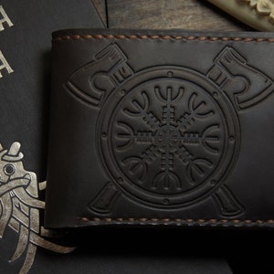 May include: Dark brown leather wallet with embossed Viking axe and shield design. The wallet features a bi-fold design with visible stitching. A book with silver text and a Viking illustration is in the background.
