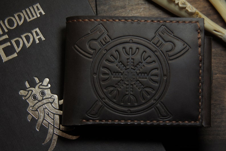 May include: Dark brown leather wallet with embossed Viking axe and shield design. The wallet features a bi-fold design with visible stitching. A book with silver text and a Viking illustration is in the background.