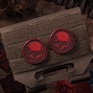 Puede incluir: Dos parches circulares con un diseño de calavera en rojo oscuro con un detalle prominente en la frente, sujetos a un panel táctico MOLLE rectangular de color verde oliva. Los parches tienen un borde rojo oscuro y están centrados en la tela.