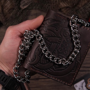 May include: A brown leather wallet with a decorative embossed design and a silver chain. The wallet is held in a hand, and the chain is draped over it. The wallet appears to be new.