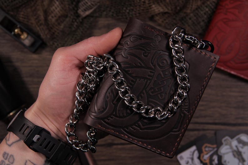 May include: A brown leather wallet with a decorative embossed design and a silver chain. The wallet is held in a hand, and the chain is draped over it. The wallet appears to be new.
