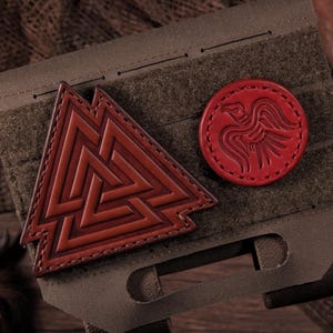 May include: A tactical olive drab MOLLE panel featuring two leather patches. One patch is a brown triangular Valknut symbol, and the other is a circular red patch embossed with a stylised raven design. The patches are attached via hook-and-loop backing.