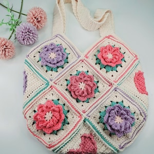 Puede incluir: Una bolsa de mano de crochet con fondo crema y diseños florales coloridos. La bolsa tiene una sola correa y está hecha con una variedad de puntos.