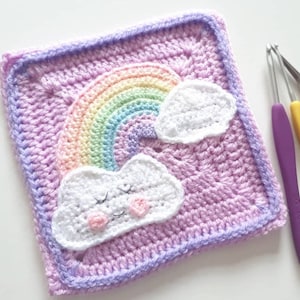 Pode incluir: Um quadrado de crochê roxo com um arco-íris e duas nuvens brancas. As nuvens têm olhos bordados e bochechas cor-de-rosa.