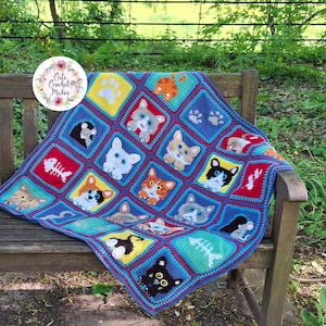 May include: A handmade crochet blanket featuring a grid of colorful squares. Each square displays a different cat face, paw print, or fish skeleton motif in various colors including yellow, red, blue, and green with a circular logo in the corner.