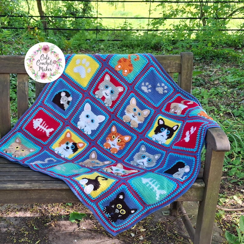 May include: A handmade crochet blanket featuring a grid of colorful squares. Each square displays a different cat face, paw print, or fish skeleton motif in various colors including yellow, red, blue, and green with a circular logo in the corner.