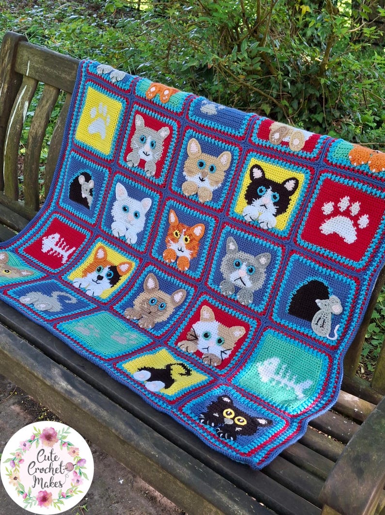 May include: A multi-colored crochet blanket featuring a grid of square panels with various cat faces, paw prints, fish skeletons, and mice. The squares are bordered in blue and red, showcasing diverse feline expressions and animal-themed motifs.