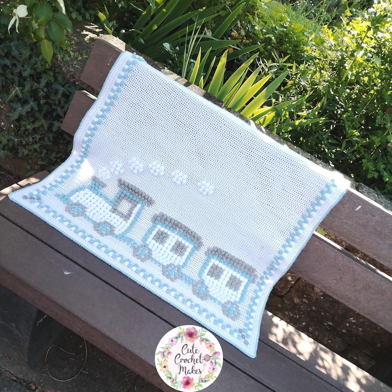 May include: A white crochet baby blanket with a blue and gray train design. The blanket has a blue and white border. The blanket is on a wooden bench.
