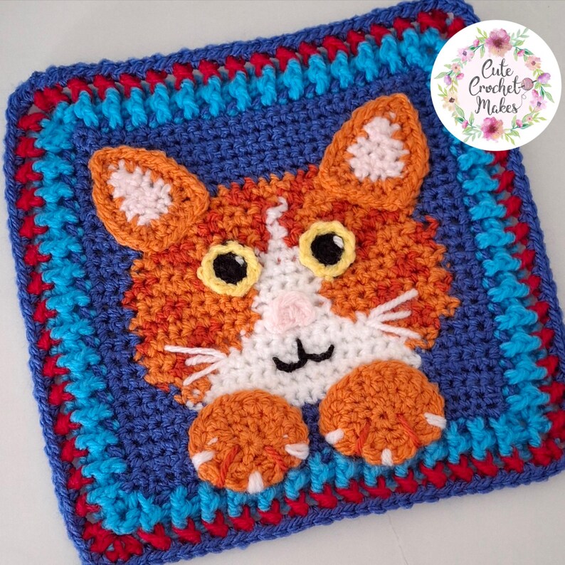 May include: A square crochet granny square featuring an orange and white cat face with yellow eyes, a pink nose, and two orange paws. The border is composed of blue, red, and light blue yarn. A circular logo in the corner reads Cute Crochet Makes.