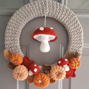 Op de afbeelding: Een handgemaakte gehaakte krans met een lichtgrijze basis. De krans is versierd met oranje pompoenen, rood-witte paddenstoelen en herfstbladeren. Een rood-witte paddenstoel hangt in het midden van de bovenkant.