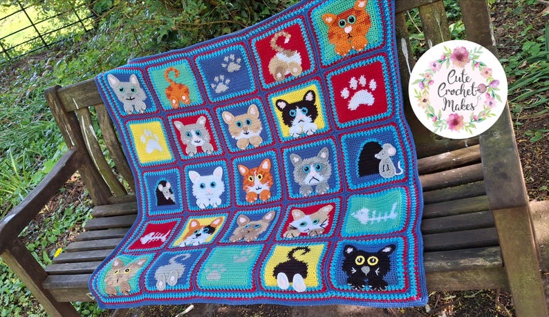 May include: A handmade crochet blanket featuring a grid of colorful squares, each displaying various cat-themed designs including cat faces, paw prints, fish skeletons, and mice. The blanket has a blue border and includes a circular logo reading Cute Crochet Makes.