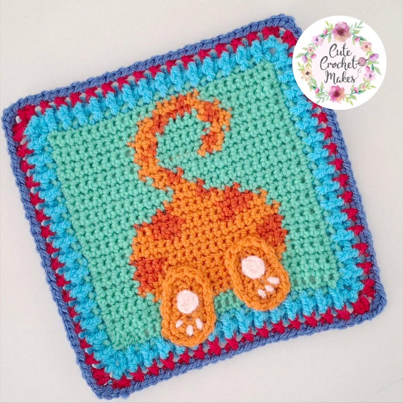 May include: A square crochet granny square featuring a central orange cat rear end motif with a raised tail and two paws on a light teal background. The border consists of alternating rows of bright blue, magenta, and dark blue yarn stitches.