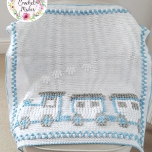 May include: A white crochet baby blanket with a blue train design. The train is made of white and gray bobbles and has three cars. There are white clouds above the train. The blanket has a blue and white border.