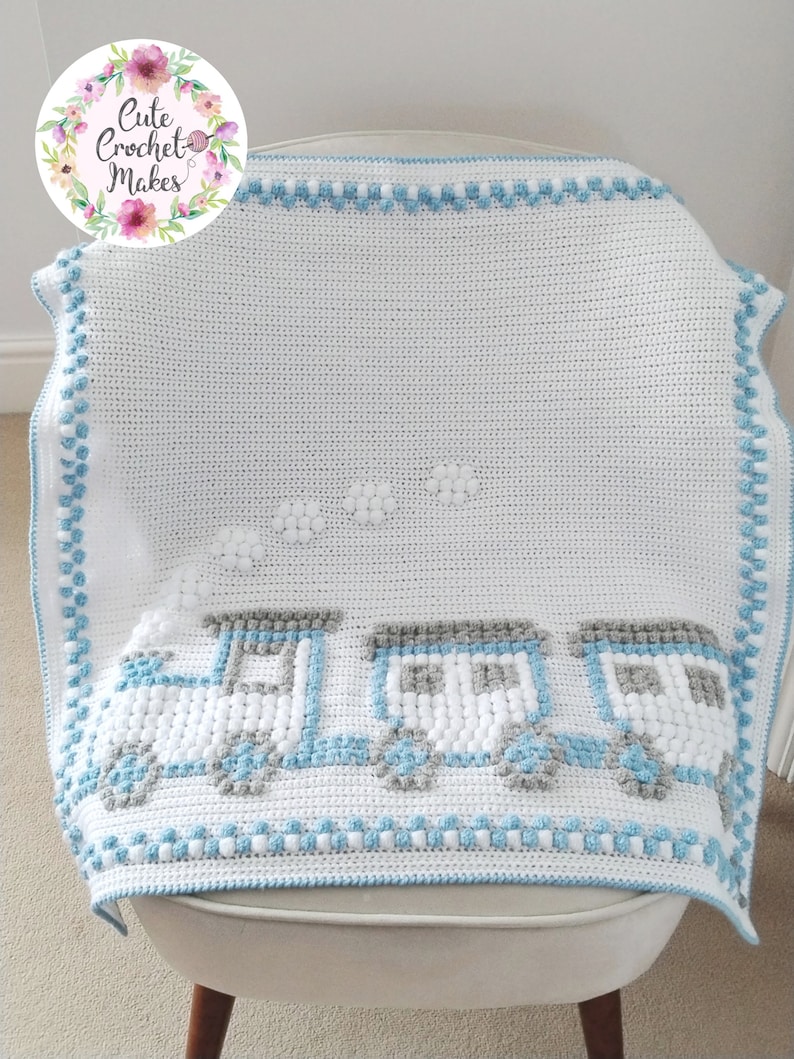 May include: A white crochet baby blanket with a blue train design. The train is made of white and gray bobbles and has three cars. There are white clouds above the train. The blanket has a blue and white border.