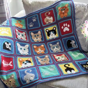 May include: A handmade crochet blanket featuring a grid of colorful squares. Each square displays various cat faces, cat tails, paw prints, a fish skeleton, and a mouse. The blanket is bordered in blue and includes a floral logo reading Cute Crochet Makes.