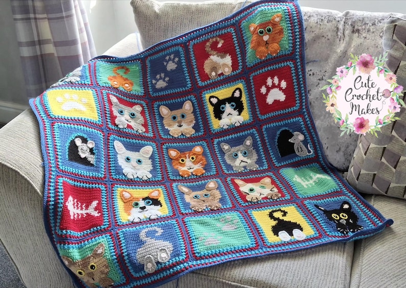 May include: A handmade crochet blanket featuring a grid of colorful squares. Each square displays various cat faces, cat tails, paw prints, a fish skeleton, and a mouse. The blanket is bordered in blue and includes a floral logo reading Cute Crochet Makes.