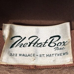 May include: A white label with brown trim and the text "The Hat Box Inc. 323 Wallace - St. Matthews" on a brown felt hat.