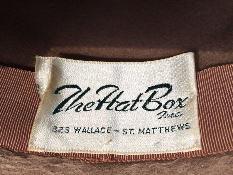 May include: A white label with brown trim and the text "The Hat Box Inc. 323 Wallace - St. Matthews" on a brown felt hat.