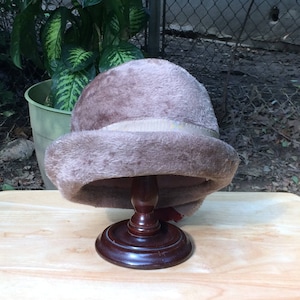 May include: A brown faux fur hat with a satin ribbon band. The hat is displayed on a wooden hat stand.